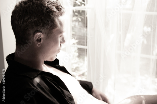 Black and white profile of sexy man in his underwear sitting in front of window and covered in bright sunshine.