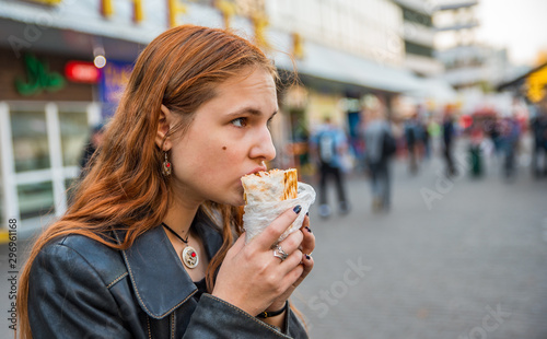 portrait of young teenager redhead girl with long hair eating chicken shawarma on street