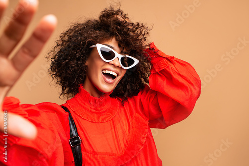 Portrait of beautiful young black woman taking selfie.Portrait of a pretty young afro american woman in retro style clothes smiling while standing and taking a selfie isolated over beige. photo