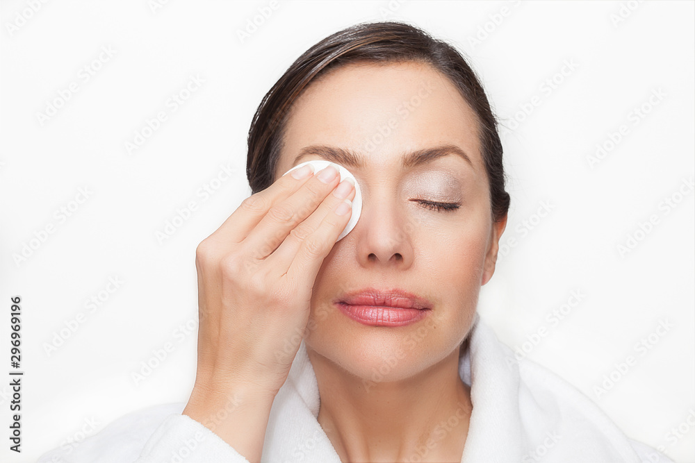 Beautiful woman removing her makeup using a cotton round wearing a white bathrobe over a white background Beautiful woman removing her makeup using a cotton round wearing a white bathrobe over a white background
