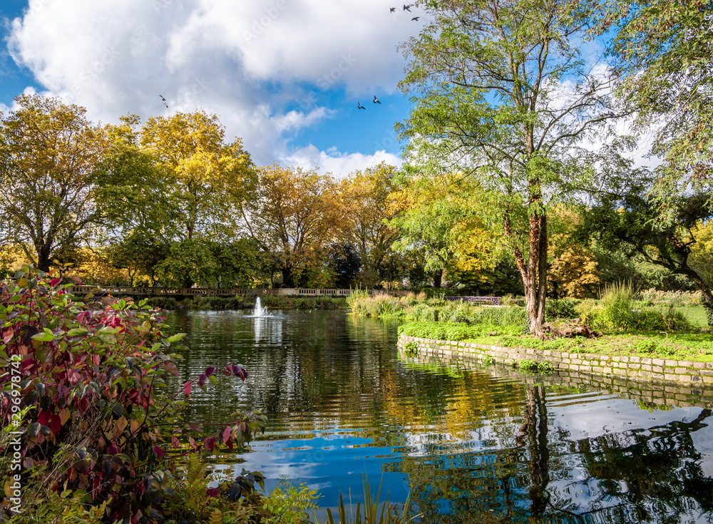 Fototapeta premium Beautiful autumn scene outdoors in Bishop public park with orange colored trees reflected in a pond in a sunny day in London