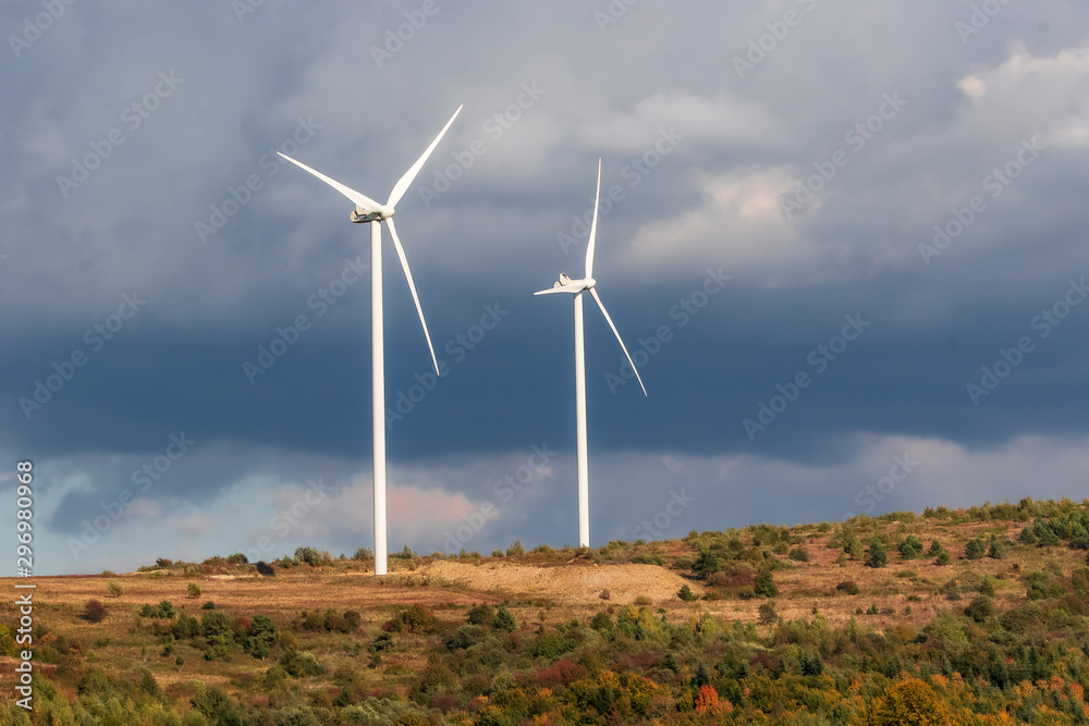 Two white wind turbines on the mountain, stormy blue sky, autumn landscape. Wind power plants. Production of alternative green renewable energy. Wind generators. Two white wind turbines on the mountain, stormy blue sky, autumn landscape. Wind power plants. Production of alternative green renewable energy. Wind generators.