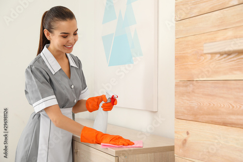 Young chambermaid wiping dust with rag in hotel room. Space for text photo