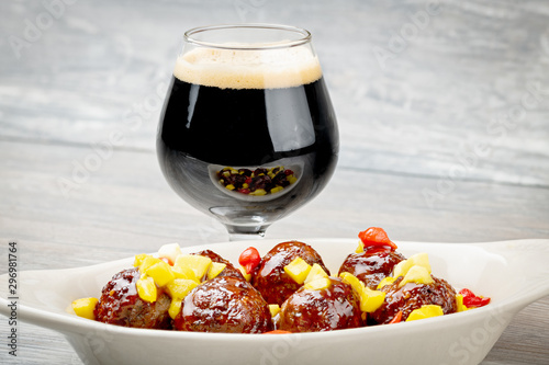 Many glistening meatballs in a white dish next to a glass of beer to be served as an appetizer