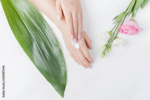 Fototapeta Naklejka Na Ścianę i Meble -  Girl puts cream on hands on white background with tropical leaves and pink flowers, top view. Concept of skin care, anti-aging frost