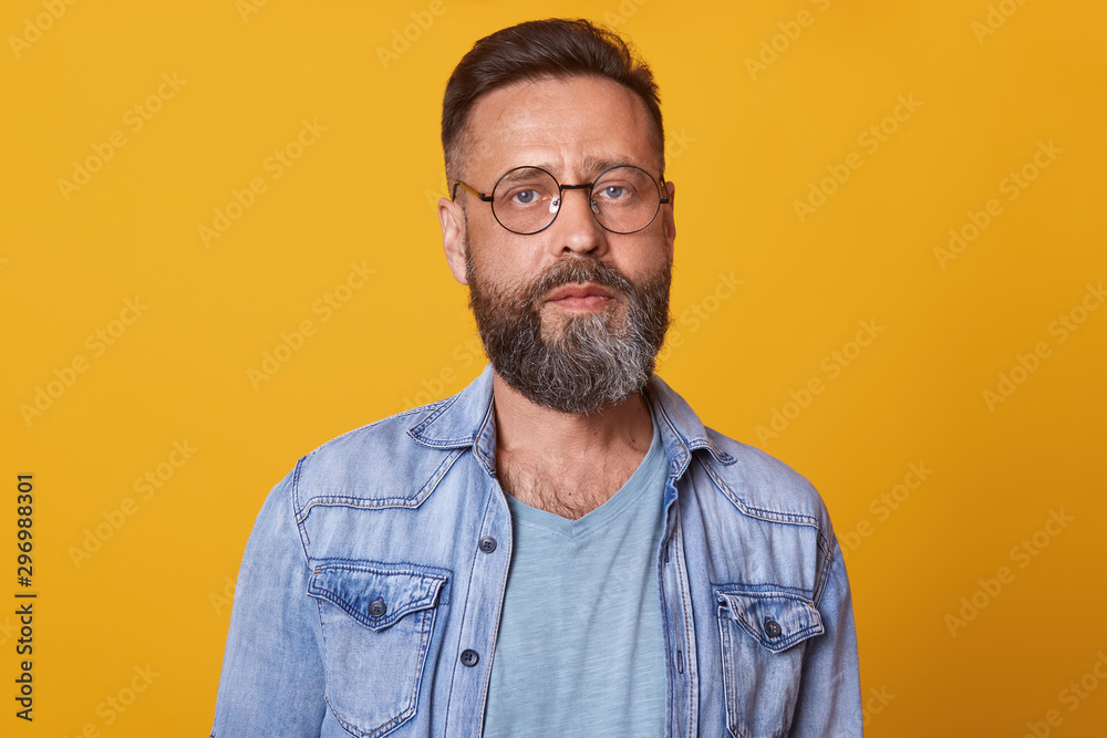 Photo of bearded man with folded hands, standing and looking directly at camera with concentrated facial expression, being in good mood, dressed stylish denim jacket and gray t shirt. People concept. Photo of bearded man with folded hands, standing and looking directly at camera with concentrated facial expression, being in good mood, dressed stylish denim jacket and gray t shirt. People concept.