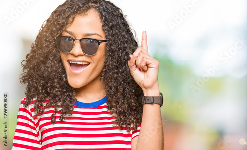 Young beautiful woman with curly hair wearing sunglasses pointing finger up with successful idea. Exited and happy. Number one.