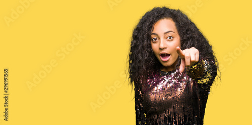 Young beautiful girl with curly hair wearing night party dress Pointing with finger surprised ahead, open mouth amazed expression, something in front