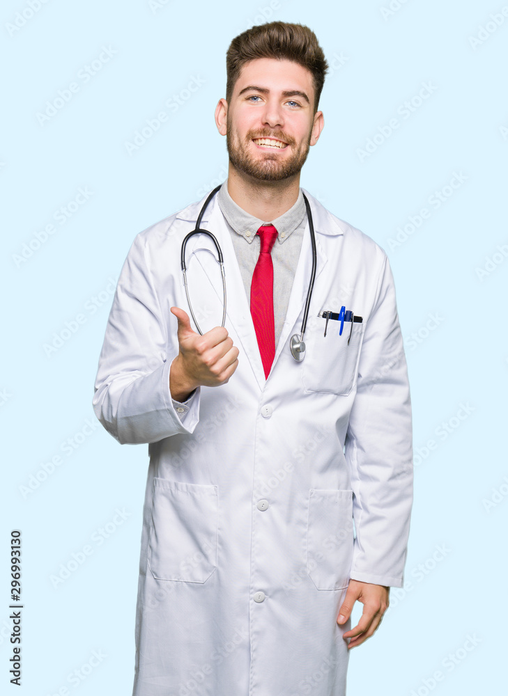 Young handsome doctor man wearing medical coat doing happy thumbs up gesture with hand. Approving expression looking at the camera showing success.