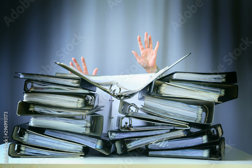 raised hands of a person who sinks behind stacks of ring binders on an office desk, concept of excessive demands and increasing work in business