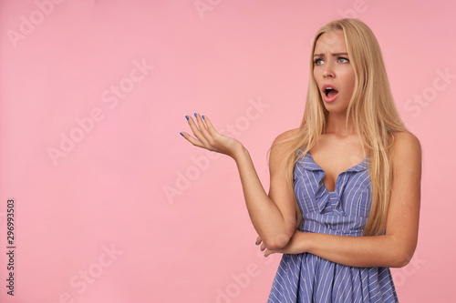 Pretty bemused young blonde woman with casual hairstyle looking aside puzzled while posing over pink background, frowning with opened mouth and raising palm