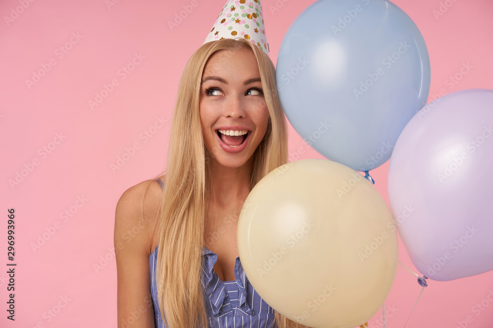 Portrait of young delighted woman with long blonde hair posing in multicolored air balloons, celebrating holiday, rejoicing nice party together with friends, standing over pink background