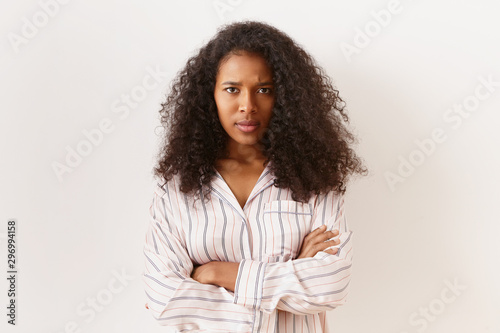 Unhappy stubborn young African American female in striped shirt posing isolated in closed posture, keeping arms crossed, frowning, expressing dissatisfaction or disagreement, feeling offended