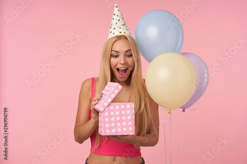 Joyful young blonde lady with long hair unpacking presents and being surprised about content, posing in multicolored air balloons over pink background, keeping her eyes and mouth wide opened © timtimphoto