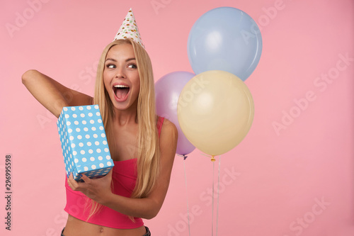 Joyful young blonde female with long hair posing over pink background while unpacking gifts, being excited and surprised to get birthday presents. People, entertainment and holiday attributes © timtimphoto