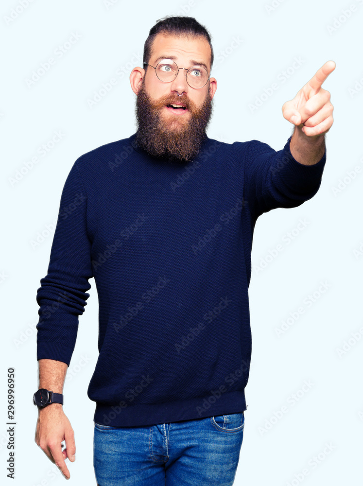 Young blond man wearing glasses and turtleneck sweater Pointing with finger surprised ahead, open mouth amazed expression, something in front Young blond man wearing glasses and turtleneck sweater Pointing with finger surprised ahead, open mouth amazed expression, something in front