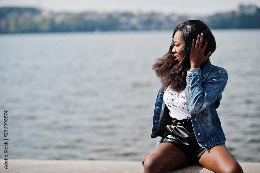 African american dark skinned slim model posed in a black shorts and jeans jacket against sea side. African american dark skinned slim model posed in a black shorts and jeans jacket against sea side.