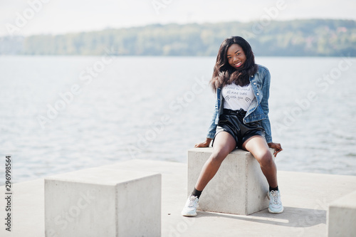 African american dark skinned slim model posed in a black shorts and jeans jacket against sea side.