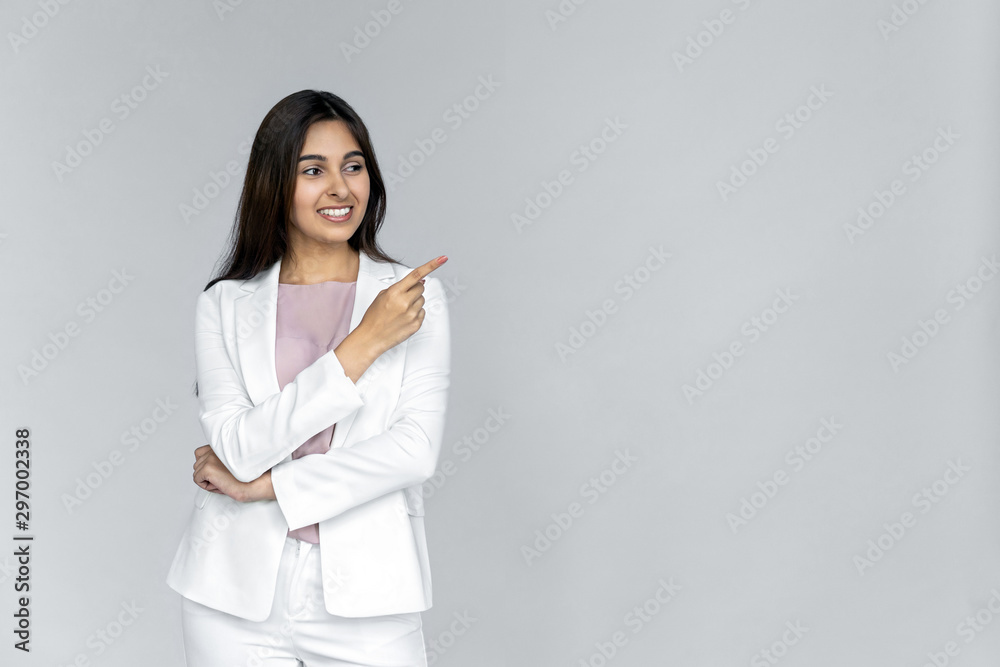 Smiling young adult indian business lady in formal white suit looking away pointing index finger aside. Happy woman professional saleswoman customer standing isolated on grey background with copyspace Smiling young adult indian business lady in formal white suit looking away pointing index finger aside. Happy woman professional saleswoman customer standing isolated on grey background with copyspace
