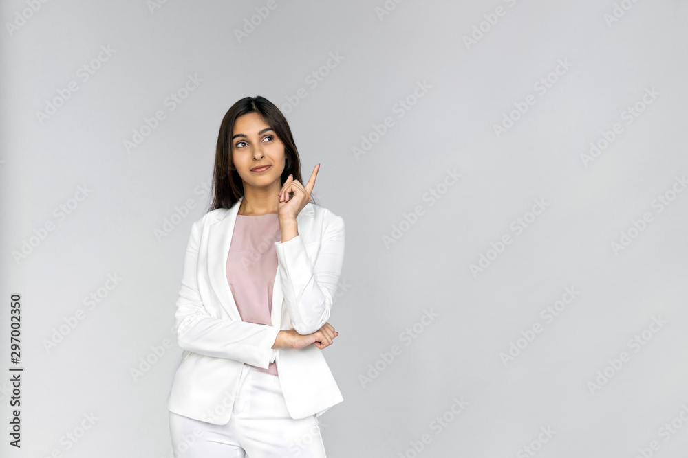 Dreamy interested young adult indian businesswoman in white formal suit wear thinking and pointing finger away at copy space. Girl smiling and standing isolated on grey background dreaming choosing Dreamy interested young adult indian businesswoman in white formal suit wear thinking and pointing finger away at copy space. Girl smiling and standing isolated on grey background dreaming choosing