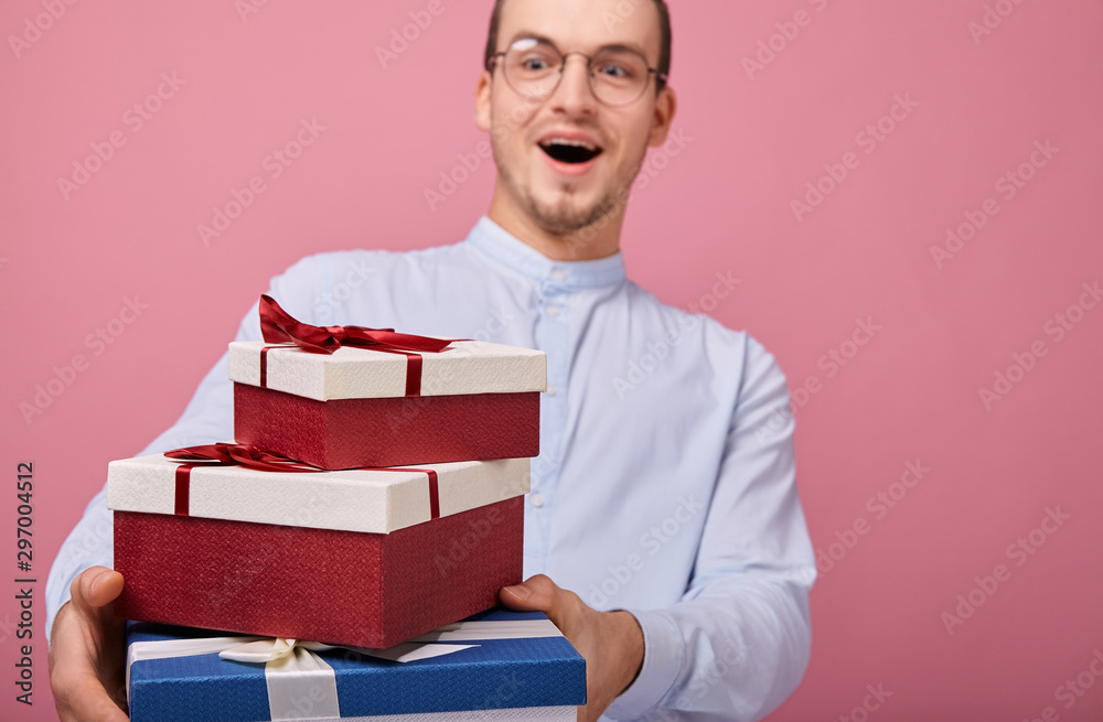 A surprised man in shirt with glasses holds gifts in a box with lid and red bow in his hands, looking on presents. Rejoicing. The guy opened mouth in surprise