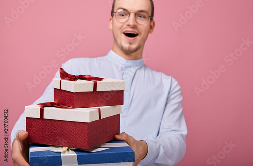 A surprised man in shirt with glasses holds gifts in a box with lid and red bow in his hands, looking on presents. Rejoicing. The guy opened mouth in surprise photo