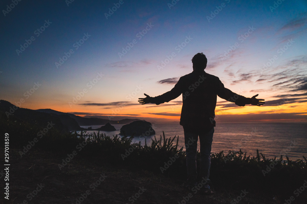 Back of a young traveler standing on on the edge of a cliff by the ocean coast watching a beautiful sunset. Arms wide open. Freedom concept