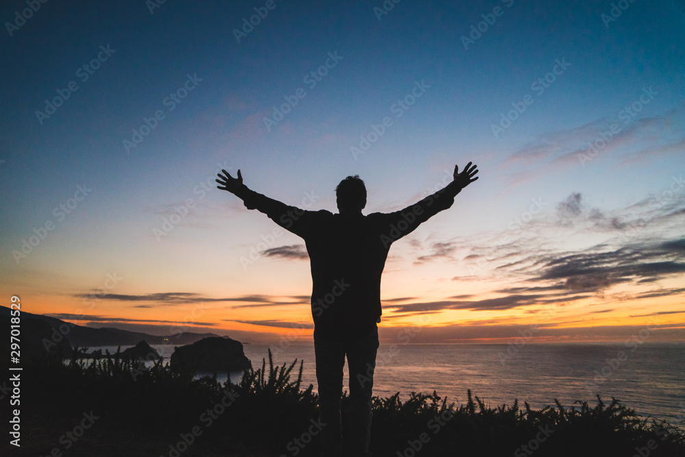 Back of a young traveler standing on on the edge of a cliff by the ocean coast watching a beautiful sunset. Arms wide open. Freedom concept