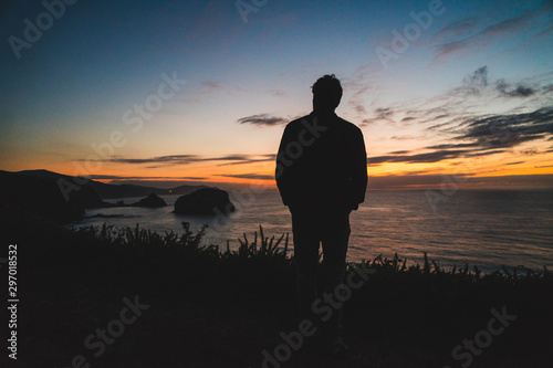 Back of a young traveler standing on on the edge of a cliff by the ocean coast watching a beautiful sunset. Freedom concept