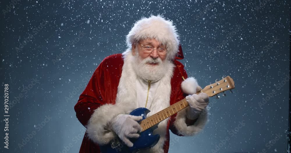 Funny santa clause is playing electric guitar and dancing while looking at camera, isolated over snowy blue background - christmas party, christmas spirit concept close up 4k footage