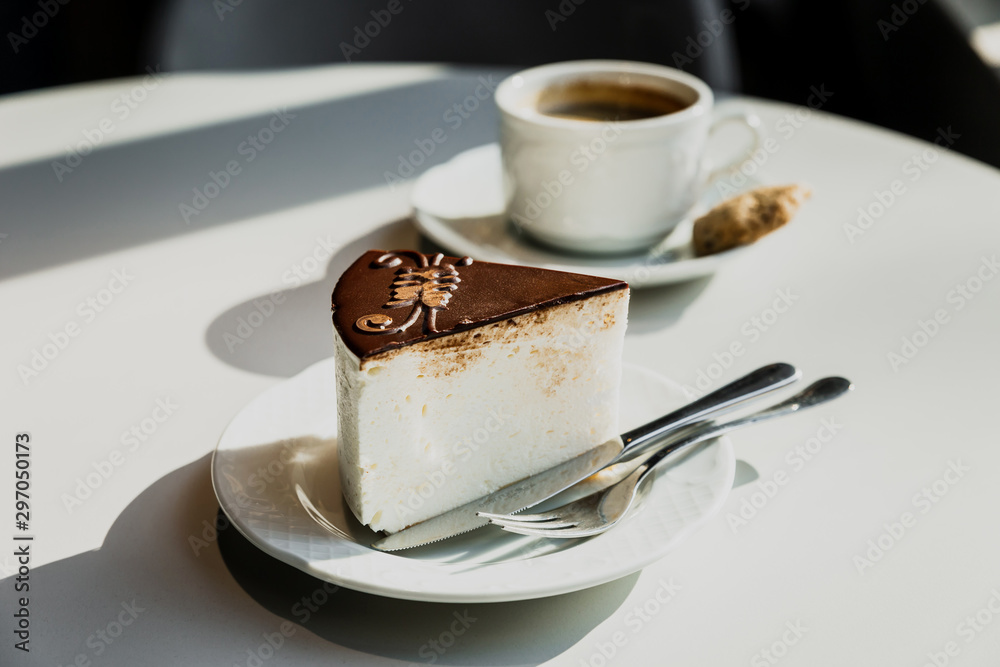 A souffle cake with chocolate on a white plate, fork, knife. A white cup of black coffee on a saucer, a cookie. Bright direct sun light. Horizontal A souffle cake with chocolate on a white plate, fork, knife. A white cup of black coffee on a saucer, a cookie. Bright direct sun light. Horizontal