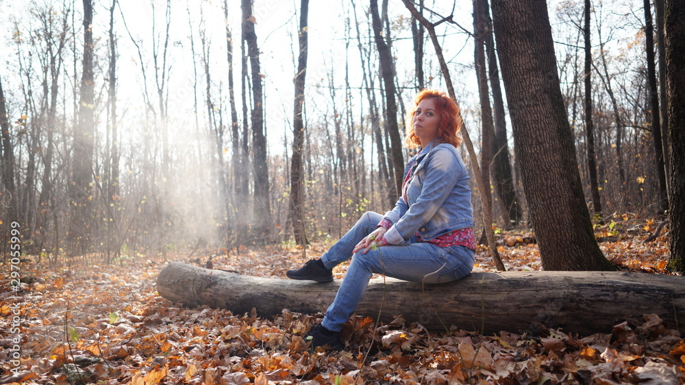 Woman sitting on log in autumn forest. Young woman with red hair in outwear sitting on fallen log in rural forest looking away in sunlight