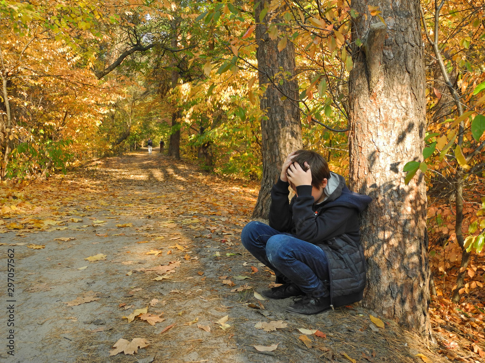 Upset problem child with head in hands sitting under tree in autumn park. Bullying, depression stress or frustration concept. Upset problem child with head in hands sitting under tree in autumn park. Bullying, depression stress or frustration concept.