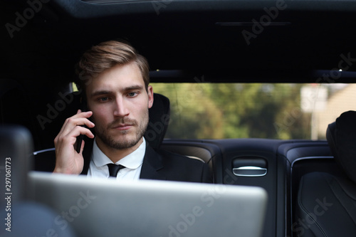 Handsome confident businessman in suit talking on smart phone and working using laptop while sitting in the car. © ty
