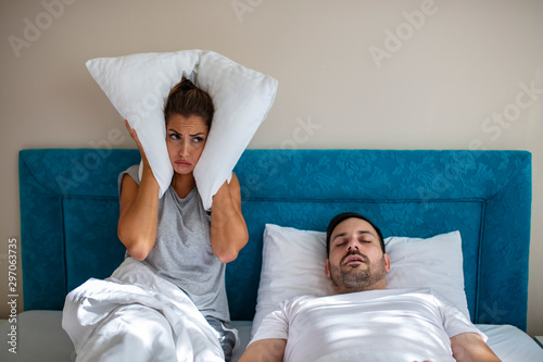 Young couple have problem with man's snoring. Heterosexual couple in bed, man sleeps and snoring with mouth open, while a tired woman irritated by snoring sitting on bed with a pillow on her head. photo