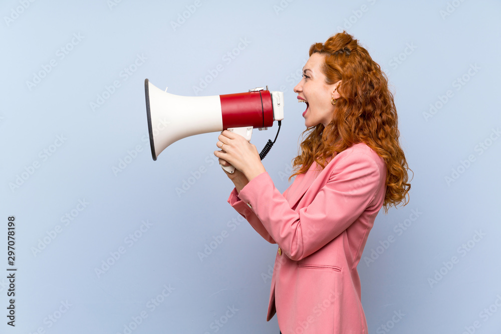 Redhead woman in suit over isolated blue wall shouting through a megaphone Redhead woman in suit over isolated blue wall shouting through a megaphone
