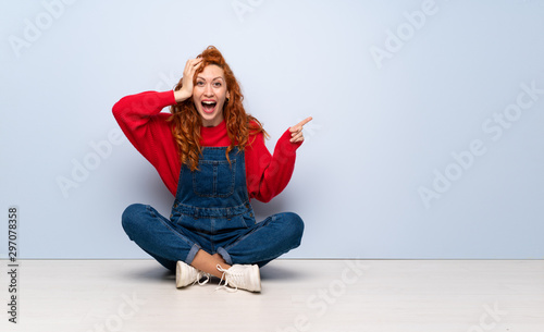 Redhead woman with overalls sitting on the floor surprised and pointing finger to the side