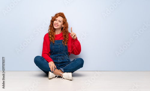 Redhead woman with overalls sitting on the floor showing and lifting a finger in sign of the best