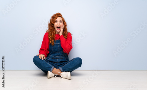 Redhead woman with overalls sitting on the floor with surprise and shocked facial expression