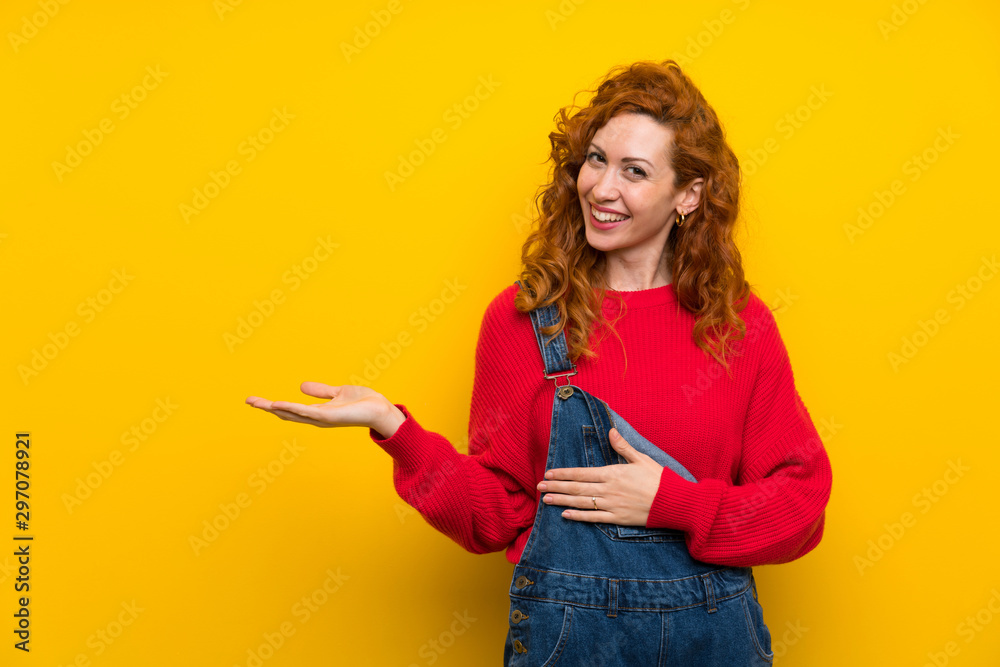 Redhead woman with overalls over isolated yellow wall holding copyspace imaginary on the palm Redhead woman with overalls over isolated yellow wall holding copyspace imaginary on the palm