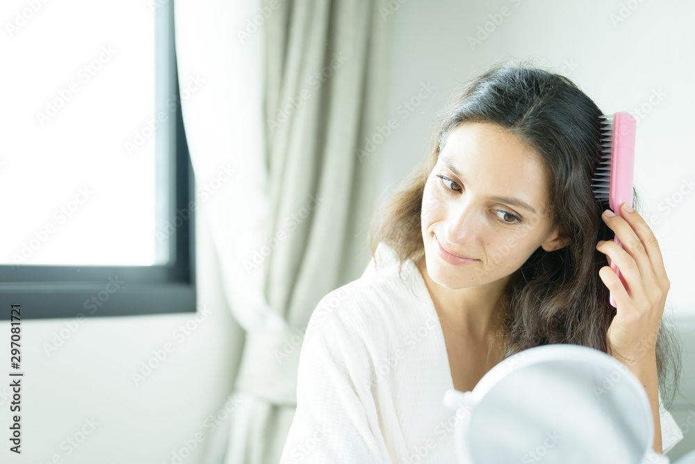 A beautiful woman wearing a towel and a white bathrobe has to look a mirror for combing hair with a pink comb and on the bed at a condominium in the morning. A beautiful woman wearing a towel and a white bathrobe has to look a mirror for combing hair with a pink comb and on the bed at a condominium in the morning.