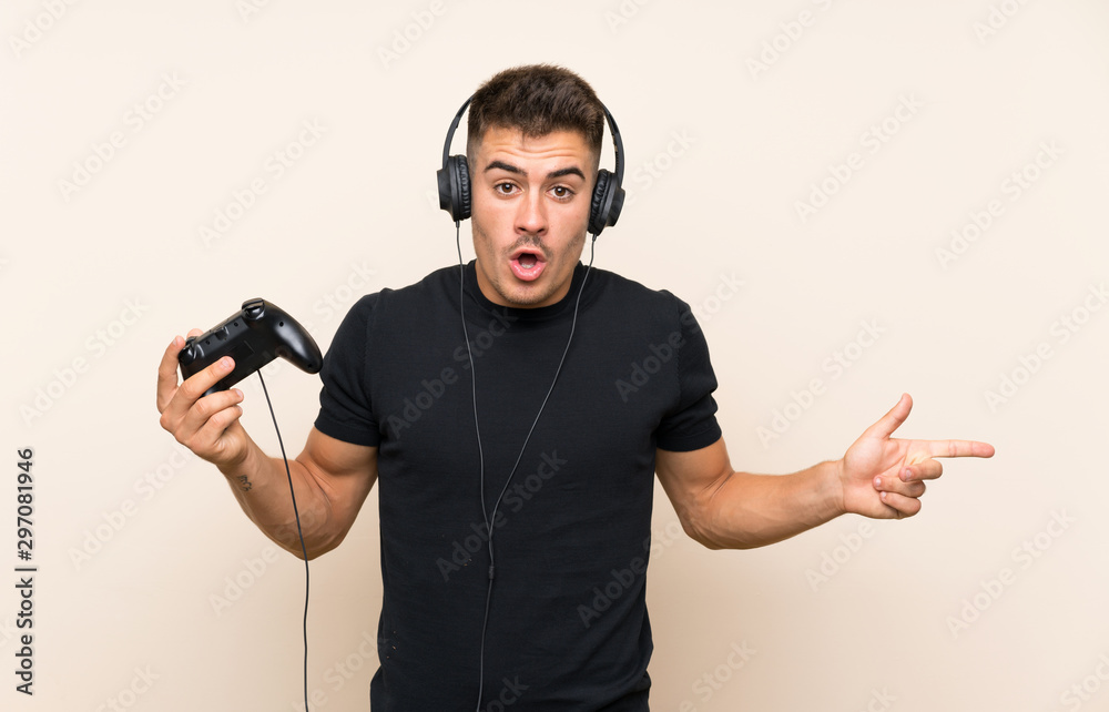 Young handsome man playing with a video game controller over isolated background surprised and pointing side Young handsome man playing with a video game controller over isolated background surprised and pointing side