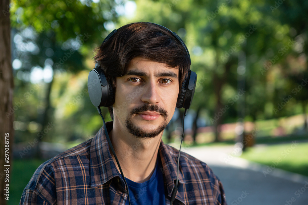 Handsome brunet bearded man in a check shirt listening to music enjoying warm day in a park. Handsome brunet bearded man in a check shirt listening to music enjoying warm day in a park.