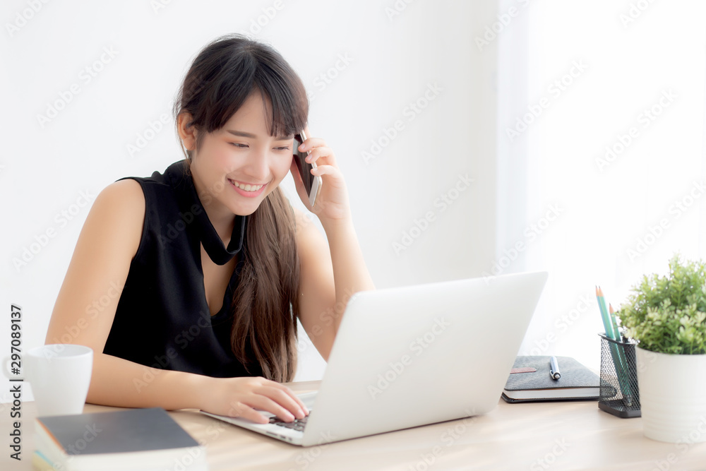 Beautiful young freelance asian woman smiling working on laptop computer and talking mobile smart phone at desk office with professional, girl using notebook, business and lifestyle concept.