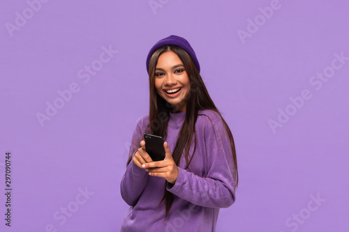 Smiling african-american girl in a hat and sweater holding smartphone isolated over purple background.