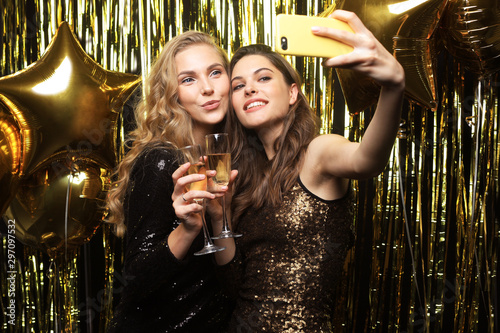 Фотография Image of two attractive girls in stylish outfit holding smartphone and taking selfie photo on gold background