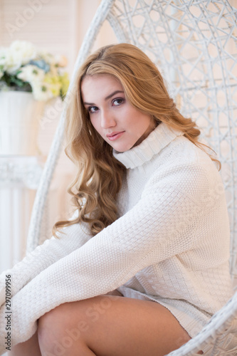 A girl in a white sweater or pullover sits in a hanging wicker chair in a cozy room in the morning.