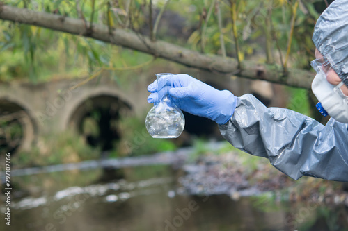 scientist, in a protective suit and mask, holds a glass flask in his hand, for taking a sample of water in the river, close-up
