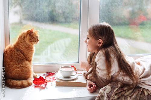 Little girl with red cat lying on window sill , reading a book and drinking cocoa. Autumn weekend with cat at home.
