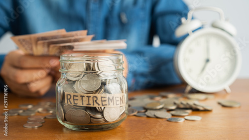 Coin in a glass bottle Image blurred background  of business people sitting  counting money and a retro white alarm clock, retirement, finance and saving money for future concept.